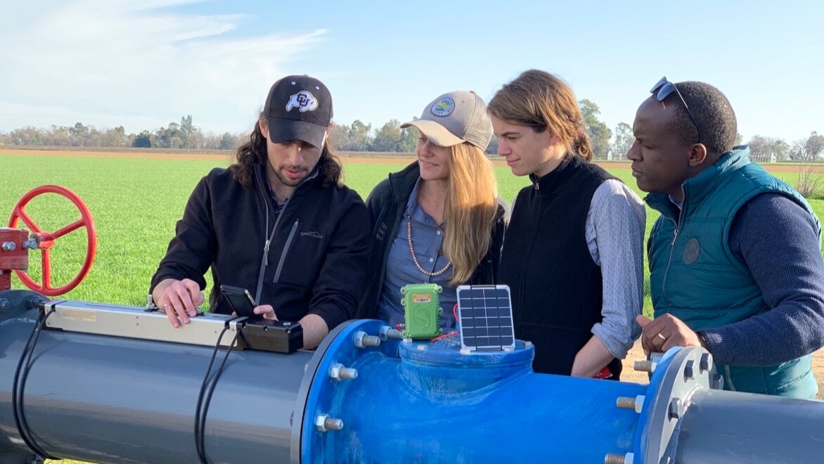 Four college students—two men and two women—standing behind and looking at a sensor mechanism on a large blue water pipe with a large green field and blue sky in the background.