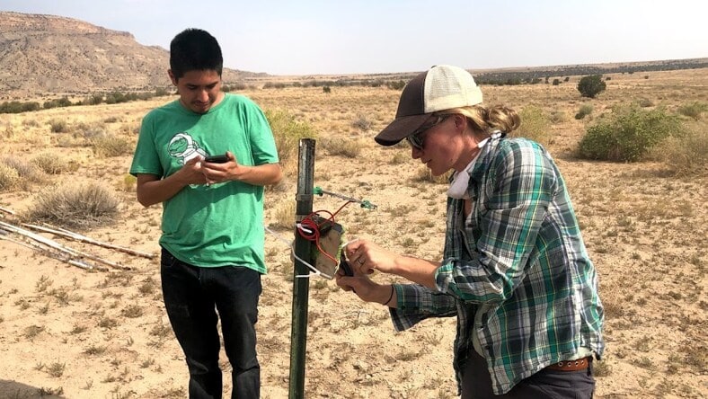 Two Mortenson Center field agents—a man (left) and woman (right)—look at a sensor on a rebar post in a large desert space with dispersed small shrubs and a mesa in the background.