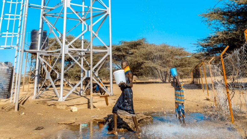 Two individuals stand under a water tower emitting water. The woman on the left is carrying a 5-gallon bucket full of water; a child on the right carries a smaller vessel, catching the water from the water tower.