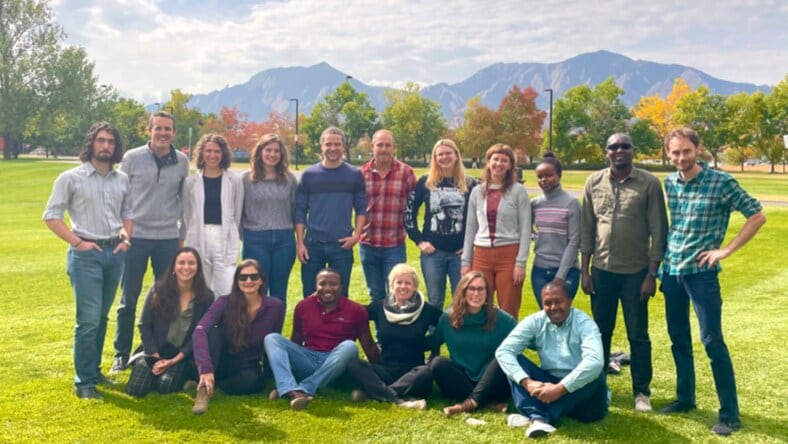 Group shot of Mortenson Center staff and students—17 individuals—sitting on a bright-green grass lawn with mountains in the background on a sunny day.