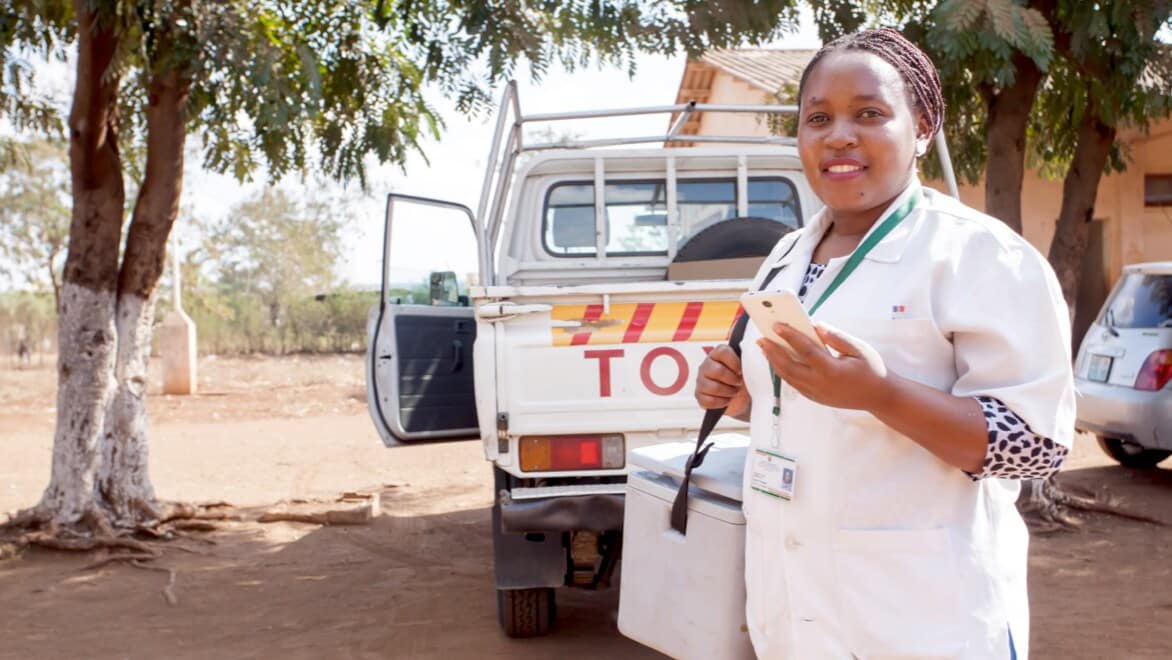 Nexleaf transport nurse—smiling, holding a smart phone, wearing white scrubs—carrying a cooler transporting Nexleaf ColdTrace technology, standing outside in front of a Toyota pickup truck with trees around it.