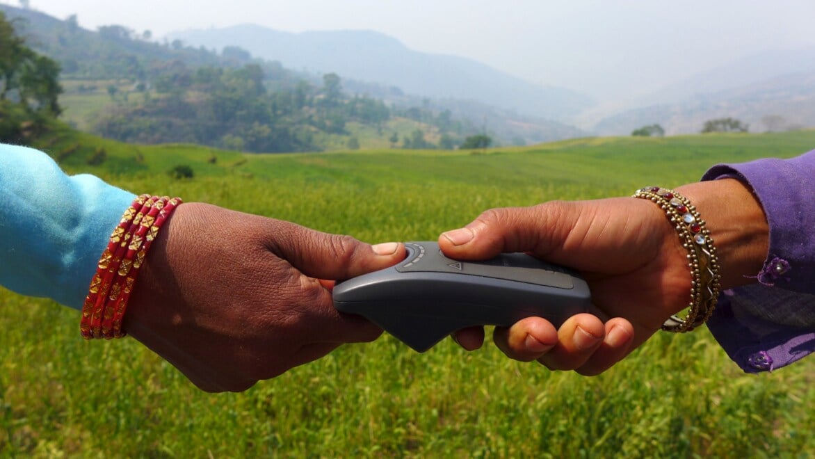 Two hands with colorful bracelets extending into the frame from the left and right borders, meeting in the middle. One is holding a Simprints device, and the other is putting a thumb on the fingerprint reader. Green grassy field and foggy mountainous landscape in the background.