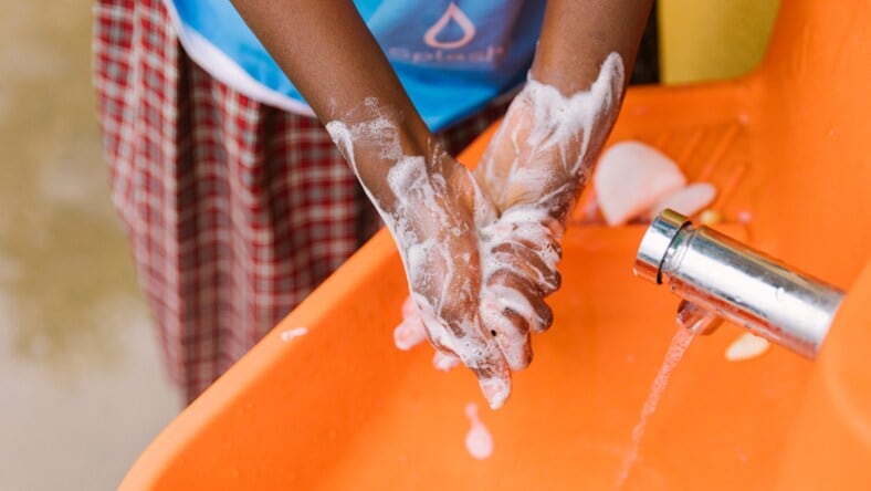 A child's hands lathered with soap, rinsing under the spigot of an orange Splash hand-washing station.