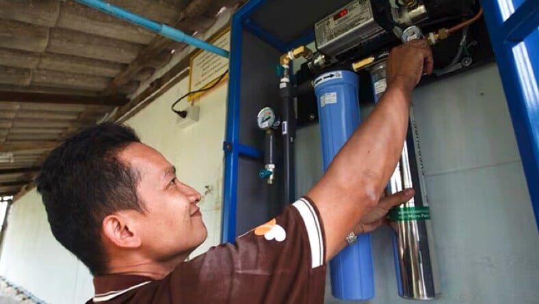 A water technician, smiling, reaching up to adjust a knob or gauge on a water filtration system.