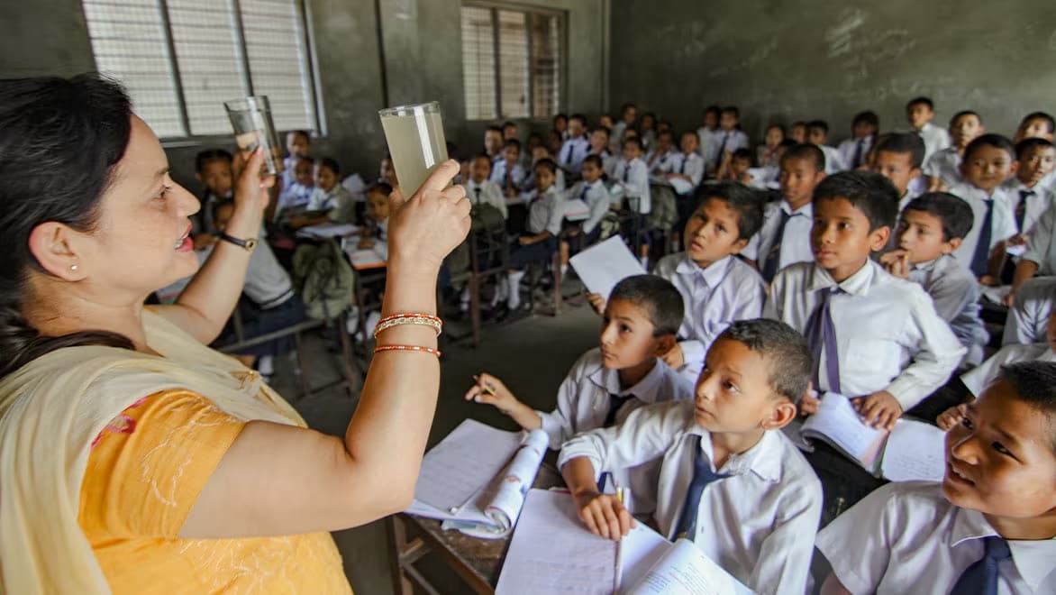 A Splash educator (woman) holds up two glasses of water in each hand—one filled with dirty/hazy water, one with clean/clear water—in front of a classroom full of schoolchildren looking along with interest.