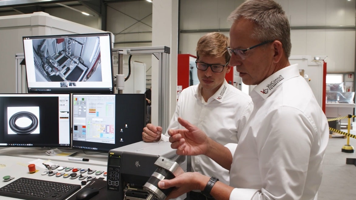 Dos hombres examinan una pieza metálica de una máquina junto a las pantallas de una máquina de inspección por rayos X