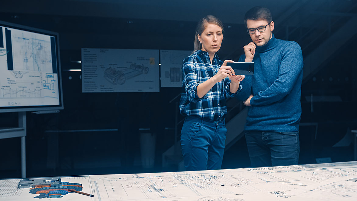 Deux jeunes développeurs étudiant les plans d’un moteur placés sur le bureau devant eux à l’aide d’un smartphone pour réalité augmentée 