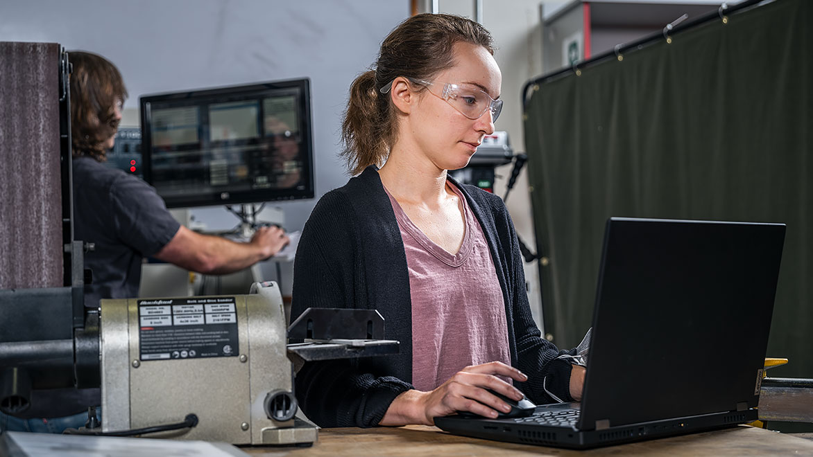 Žena v ochranných brýlích pracuje na notebooku na pracovišti.