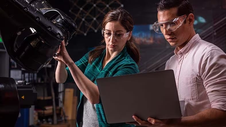 A woman and man wearing safety goggles consult a laptop computer as they work on a large machine in a robotics lab