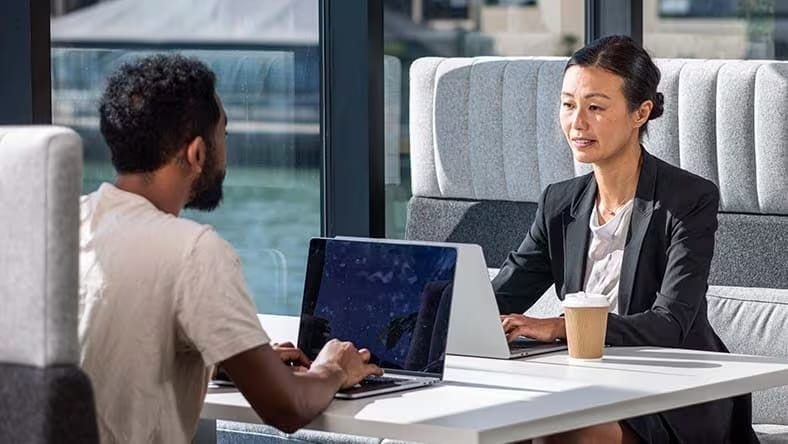 A man and woman sit across from each other at a booth, each working on their laptop computer while they collaborate