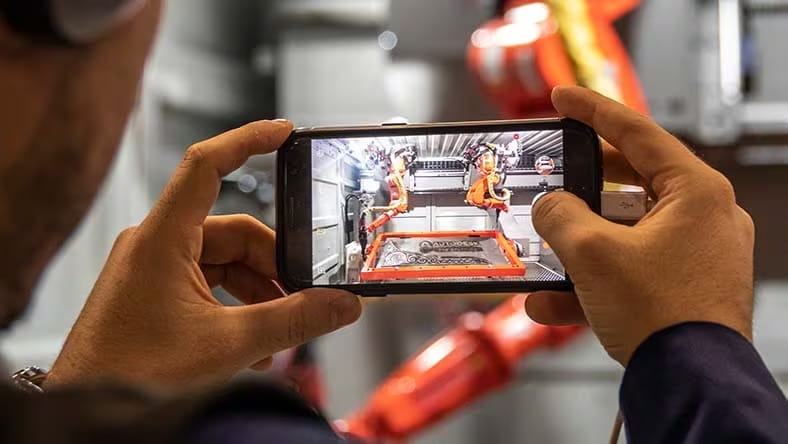 A man holds up his phone, an image of robotic arms in a factory on the screen.