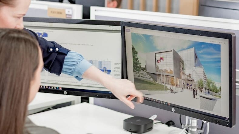 Two young women look at a desktop computer monitor displaying a 3D visualization of a building design project.