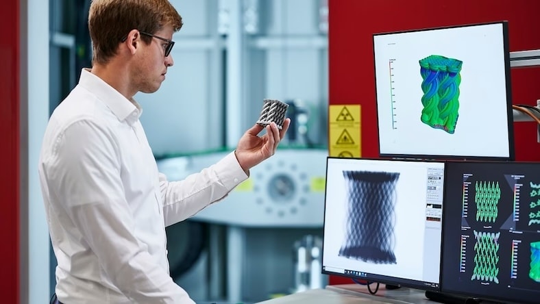 A man views images of and analyses from an X-ray inspection of the small 3D-printed object he holds in his hand.