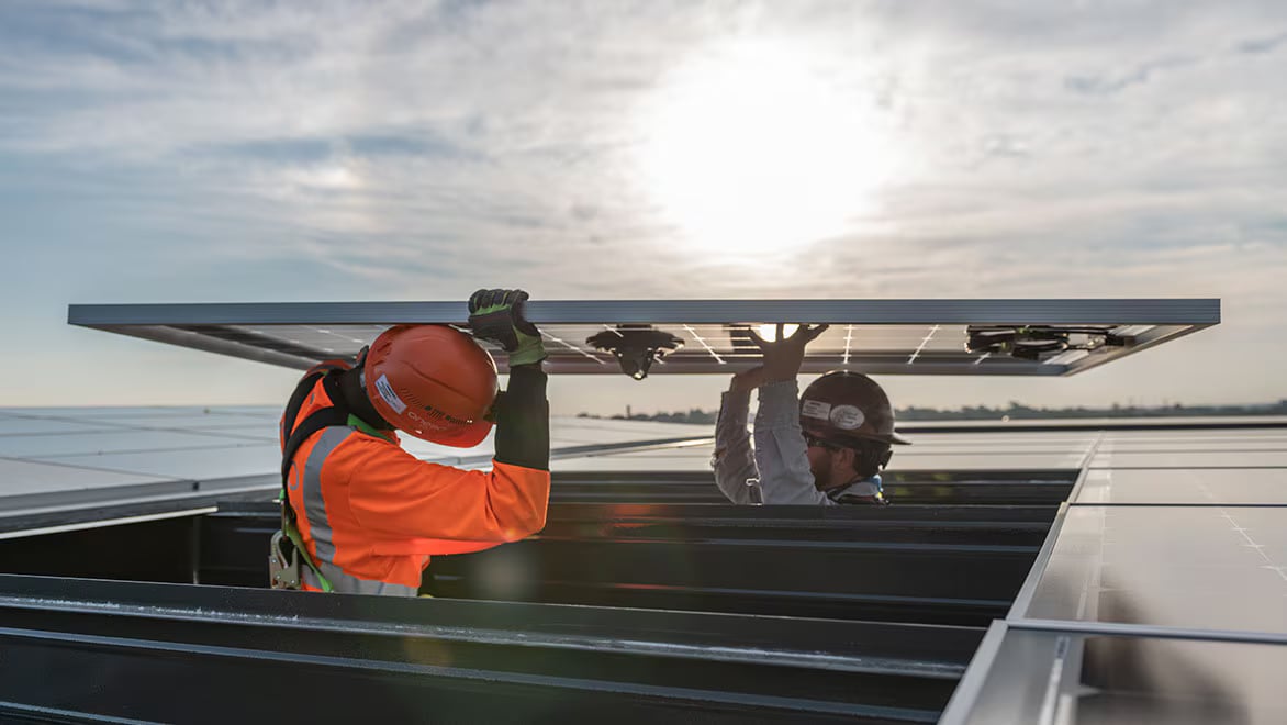 Two workers in safety gear install a solar panel on a rooftop under a bright sun. One wears an orange vest, and the other wears a gray jacket. The sky is partly cloudy, and the sun is shining brightly behind them.