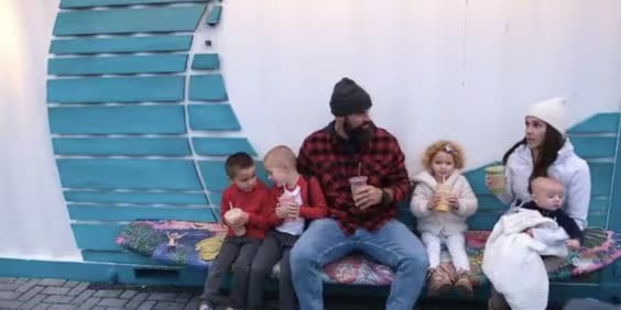 A family sits on a colorful bench against a teal and white wall. The father, wearing a beanie, holds a drink. Four children sit beside him, three with drinks. The mother, in a white hat, holds a drink and a baby wrapped in a blanket.