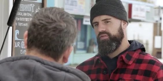 Two men are engaged in conversation outside. The man on the right has a thick beard and is wearing a black beanie and a red and black checkered flannel jacket. He appears to be listening intently. The man on the left, seen from the back, has gray hair and is wearing a dark gray jacket. In the background, there are signs and a light blue building, suggesting a casual, outdoor setting such as a market or roadside stall.