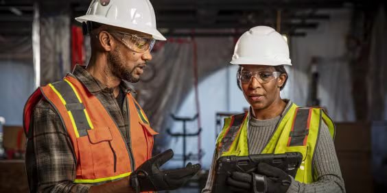 Two construction workers wearing hard hats and safety vests stand in a dimly lit worksite. One holds a tablet, and they appear to be discussing plans. The background shows construction materials and partially visible equipment.