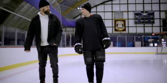 Two people wearing beanies walk on an ice rink. One person is in casual clothing, and the other is in hockey gear. They are engaged in conversation, with empty bleachers and banners visible in the background.