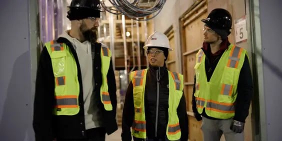 Three people wearing hard hats and reflective safety vests stand in a construction area, engaged in conversation. They are surrounded by wires and construction materials.