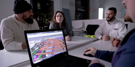 A group of four individuals are seated around a white conference table in an office setting, engaged in a discussion. The focus is on a laptop in the foreground displaying a colorful 3D architectural model. The model includes various geometric shapes in vibrant hues such as blue, orange, pink, and green, representing different components of a design. On the left, a person wearing a black beanie and a light sweater speaks while gesturing with their hands. In the center, a woman in a dark jacket attentively listens, while on the right, a man in a white shirt sits with his hands clasped. A fourth person, partially visible and out of focus on the right, operates the laptop. The background features modern office furniture and a large window.