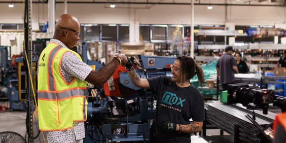 Two workers in a busy factory fist-bump. One wears a safety vest, and the other is in a Nox Innovations t-shirt, standing near machinery and stacks of materials. Other workers are visible in the background.