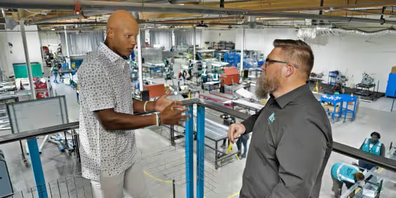 Two men are engaged in conversation on a balcony overlooking a large industrial workspace filled with machinery and workers. One man wears a patterned shirt, and the other wears a dark shirt with glasses. The workspace is bright and organized.