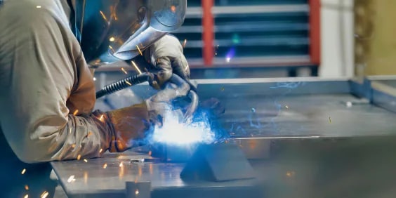 A welder in protective gear uses a welding torch, creating bright sparks and blue light on a metal surface in an industrial setting. Shelving with tools and equipment is blurred in the background.