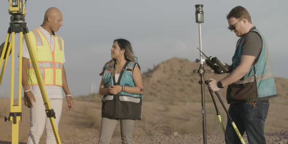 Three people conducting a land survey in an outdoor setting. Two are conversing, wearing safety vests, while the third operates surveying equipment. A tripod and a hilly landscape are visible in the background.