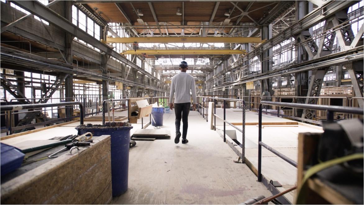 A person walks through a large, industrial warehouse with high ceilings, surrounded by metal beams and machinery. The space appears under construction or being renovated, with scattered materials and a partially assembled setting.
