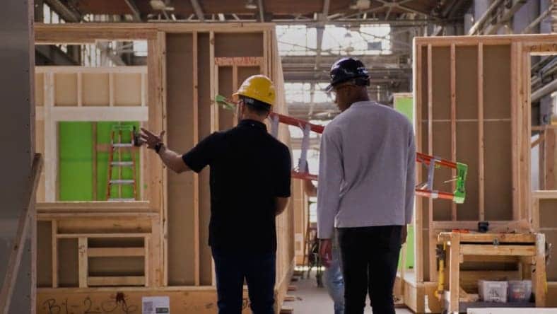 Two construction workers in hard hats stand inside an unfinished building. They are discussing plans, surrounded by wooden frames and tools. Natural light enters through large windows, highlighting the industrial space.