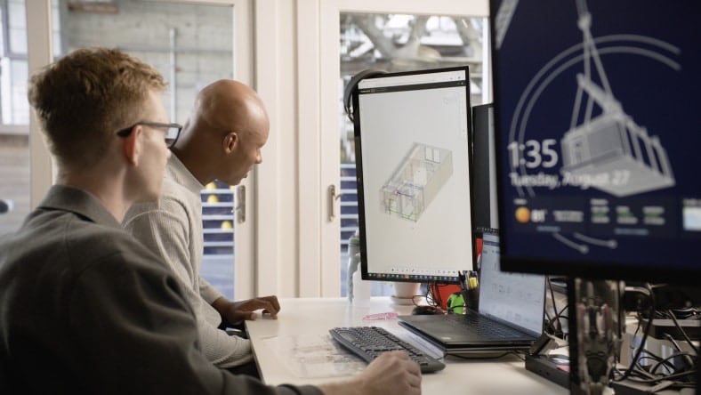 Two people at a desk look at architectural designs on a computer monitor. Another screen displays technical information, and there's a blueprint on the table.