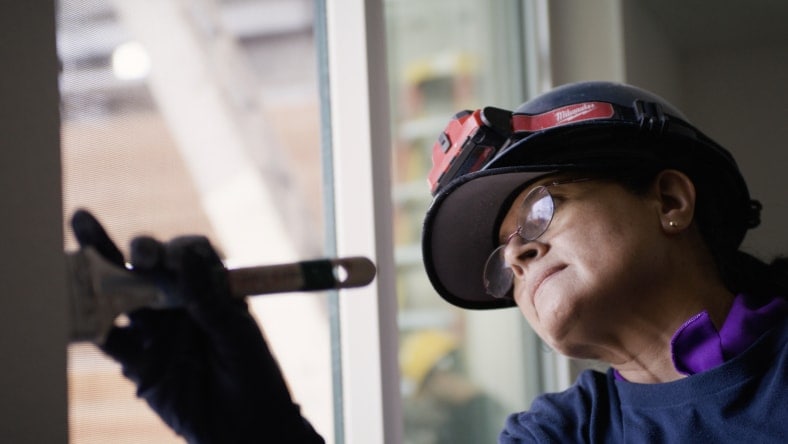 A person wearing a hard hat and glasses is concentrating while painting a window frame. They hold a paintbrush with a gloved hand, focusing intently on the task. Natural light from the window brightens their workspace.