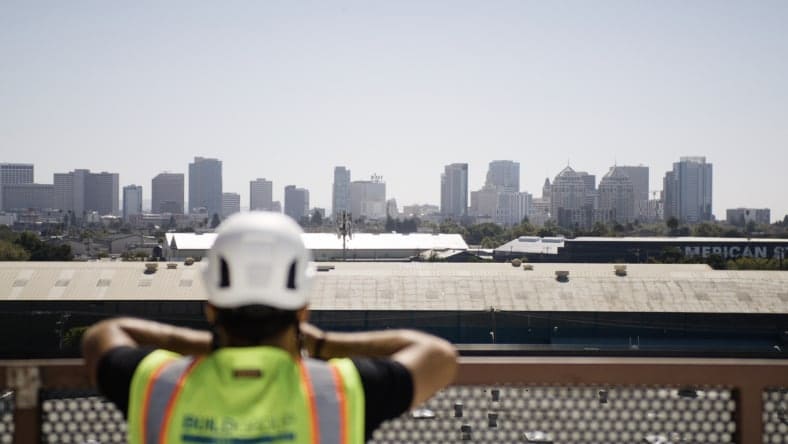 A construction worker in a safety vest and hard hat observes a city skyline from a high vantage point. The sky is clear, and the buildings in the background form a modern urban landscape.