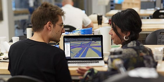 A man and a woman in an office talk while viewing a model of a highway on laptop