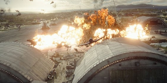 Caça voando sobre hangar com explosões geradas por computação gráfica em segundo plano