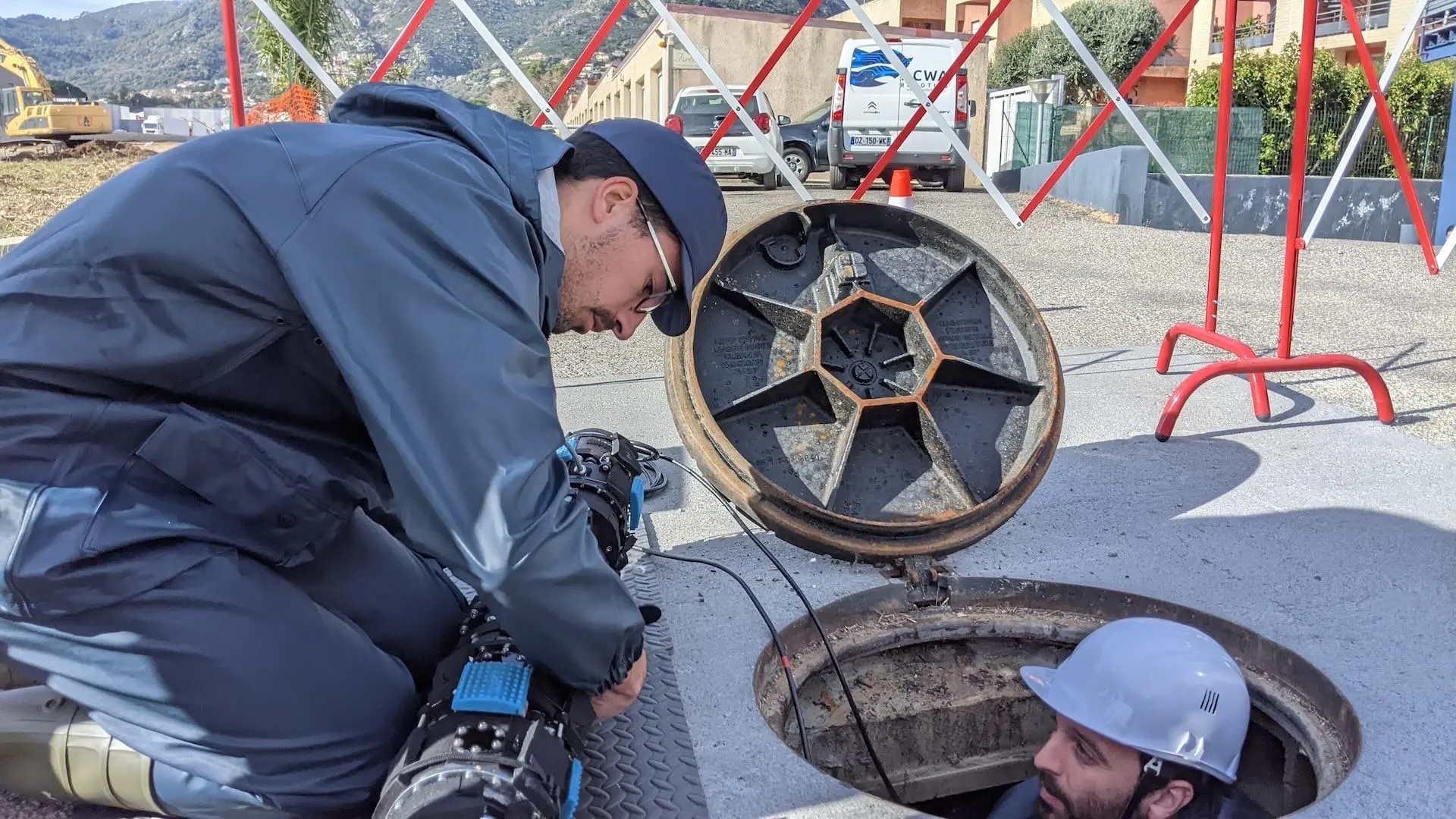 A worker with an ACWA autonomous robot kneels next to a maintenance hole in which another worker is waiting.
