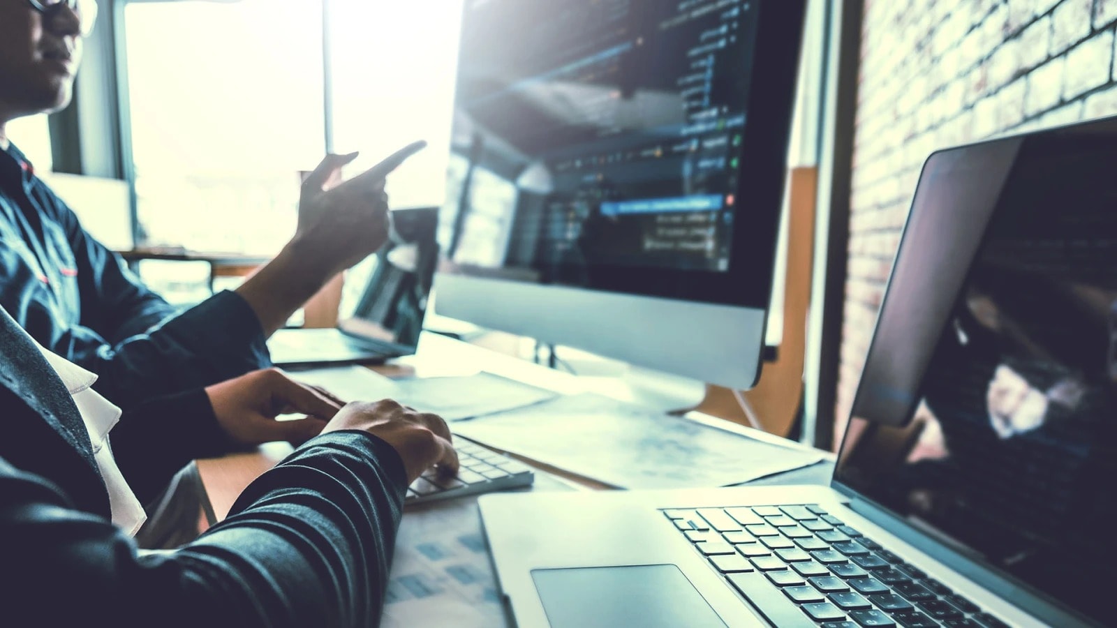 A man sits pointing at a computer workstation by a window.