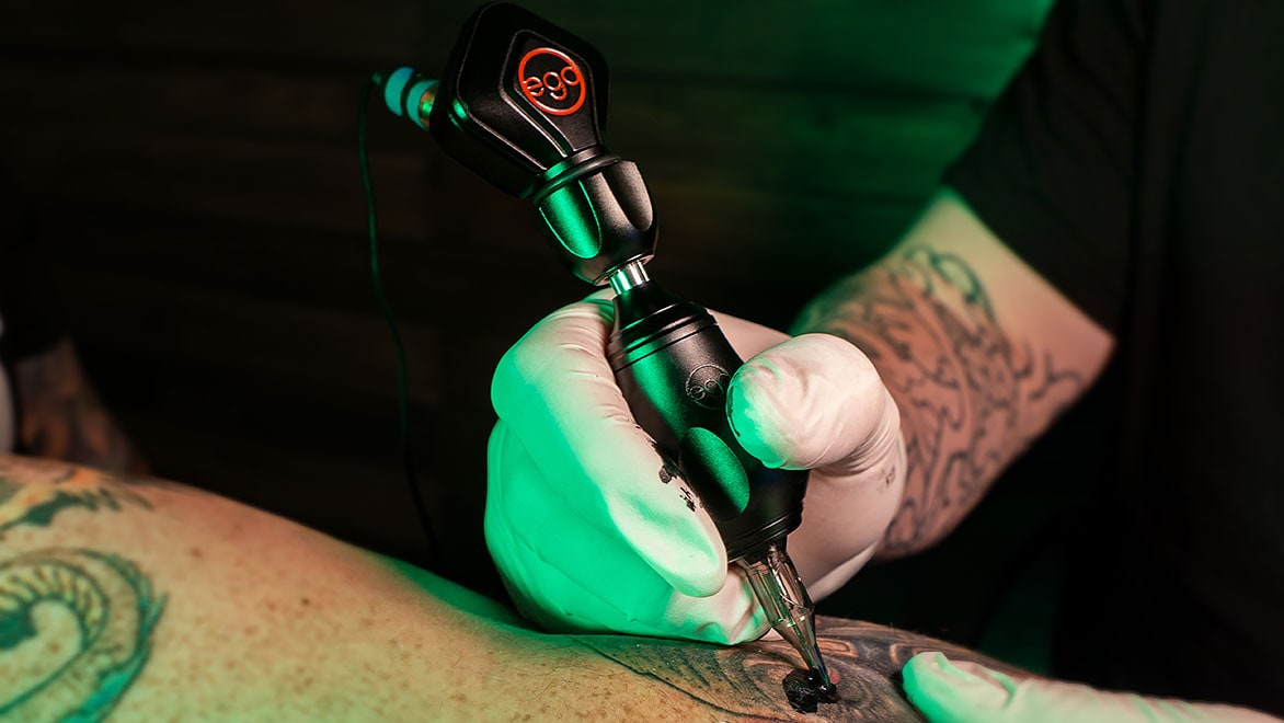 A hand in a white glove using a black Ego tattoo needle on an arm