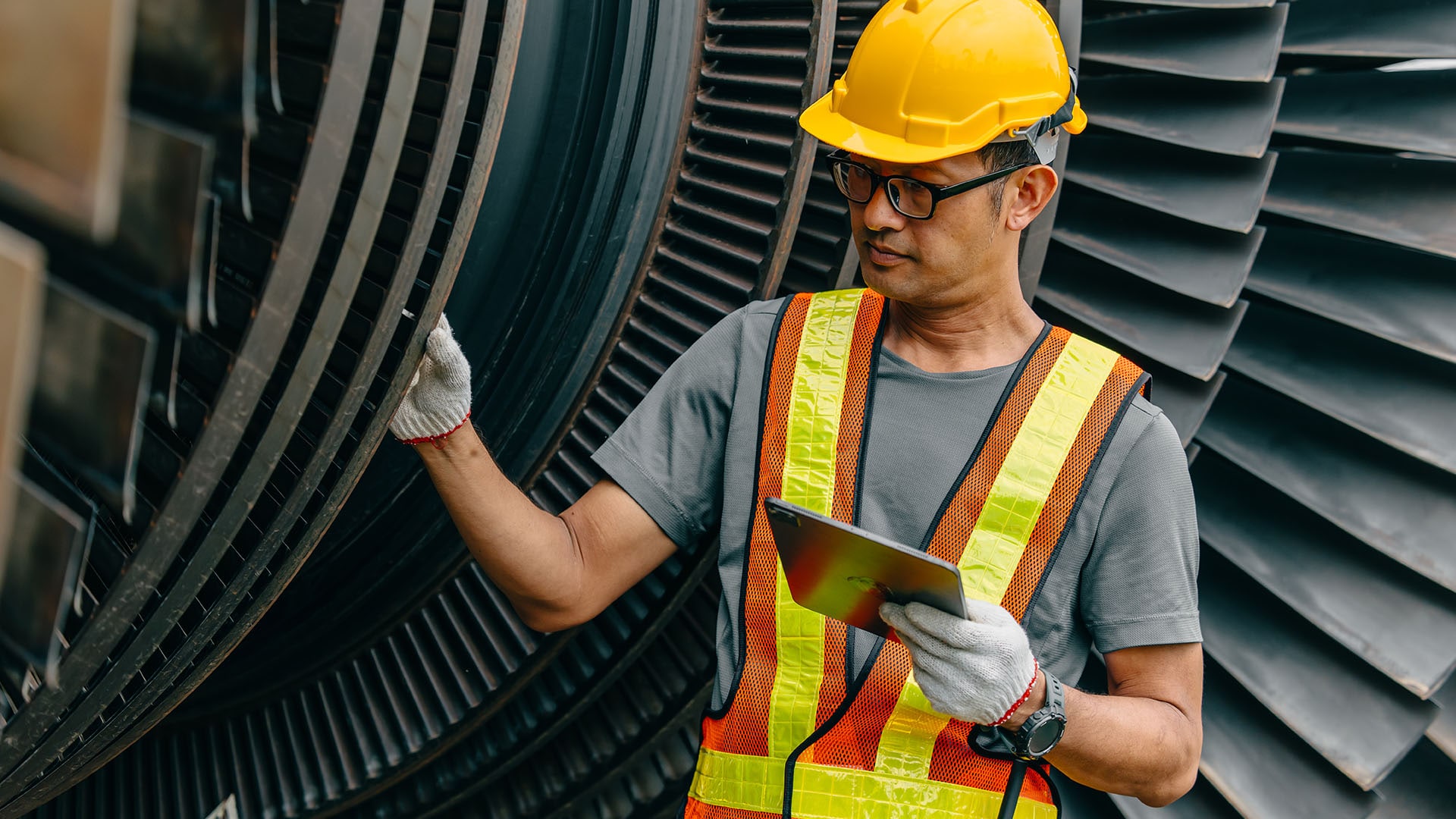Man wearing glasses inspects industrial equipment wearing high-vis vest, hard hat, and safety gloves. 