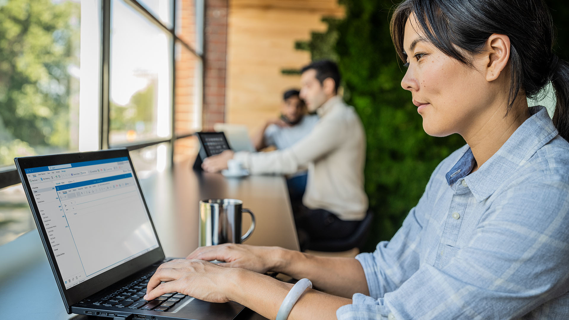 A professional woman sits in front of open laptop next to mug. Two men stand in front of open laptops in the background. 