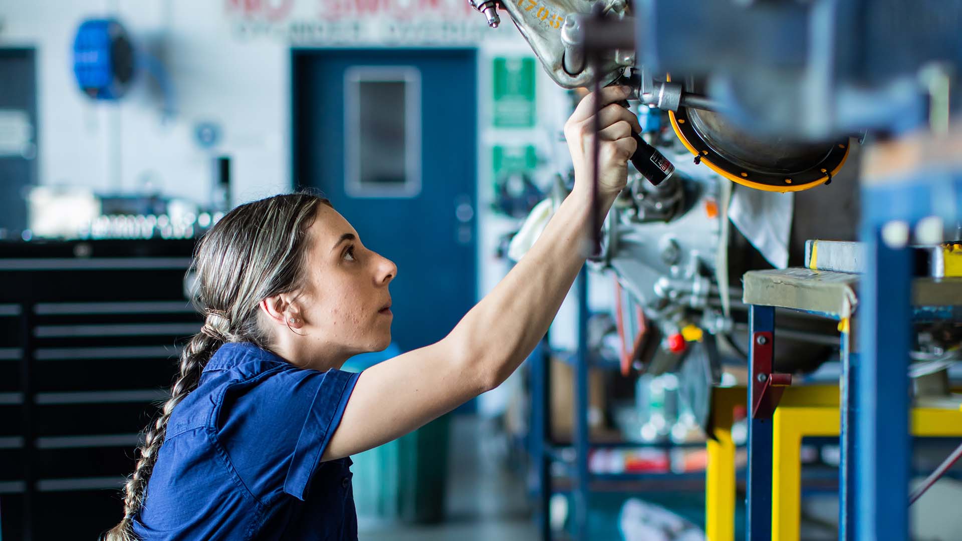Woman squats in an automotive workshop, reaching up to work on a raised vehicle. 