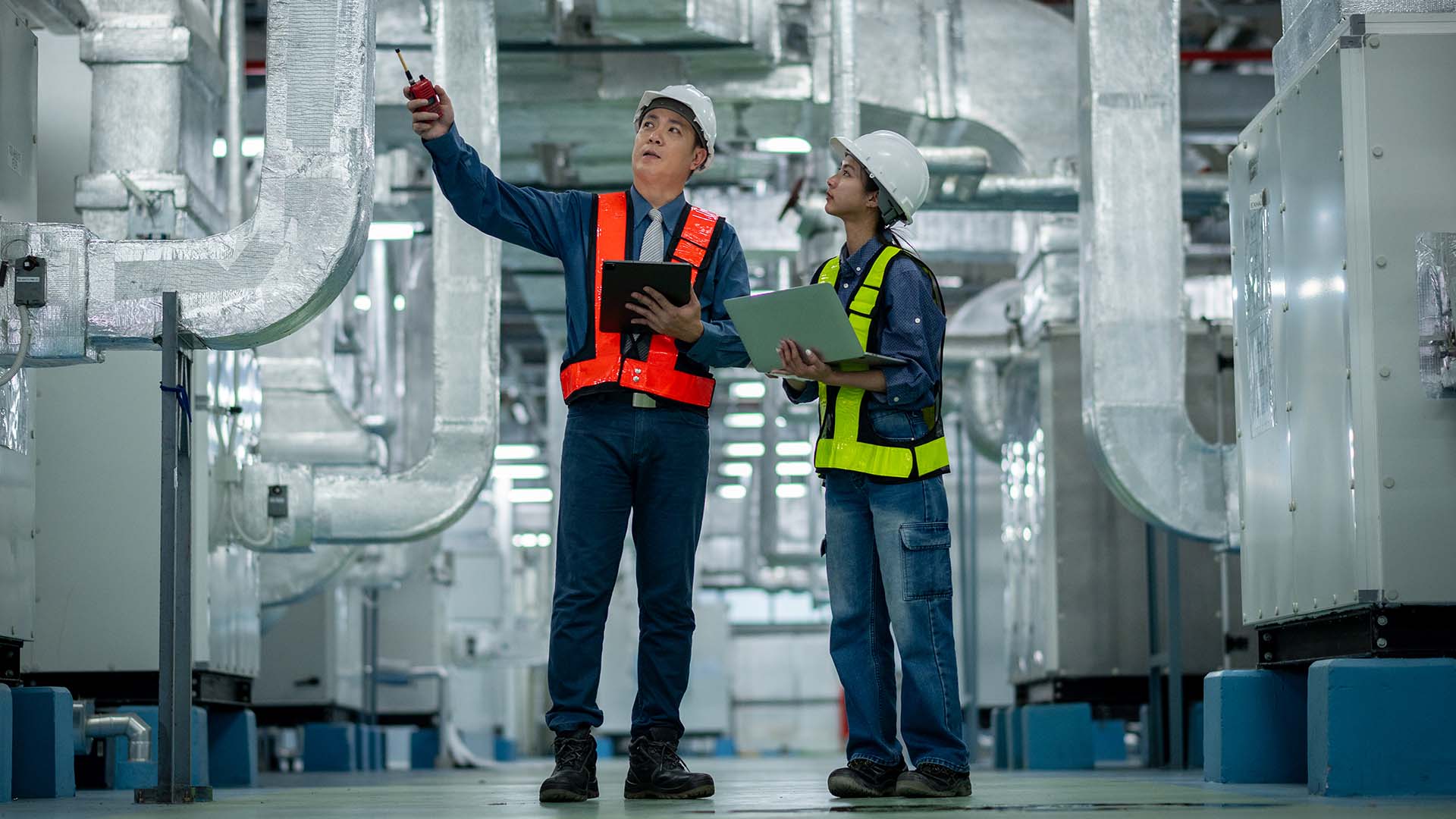 Man in high vis vest and hard hat carries tablet in factory. Woman stands next to him wearing the same gear with open laptop. 
