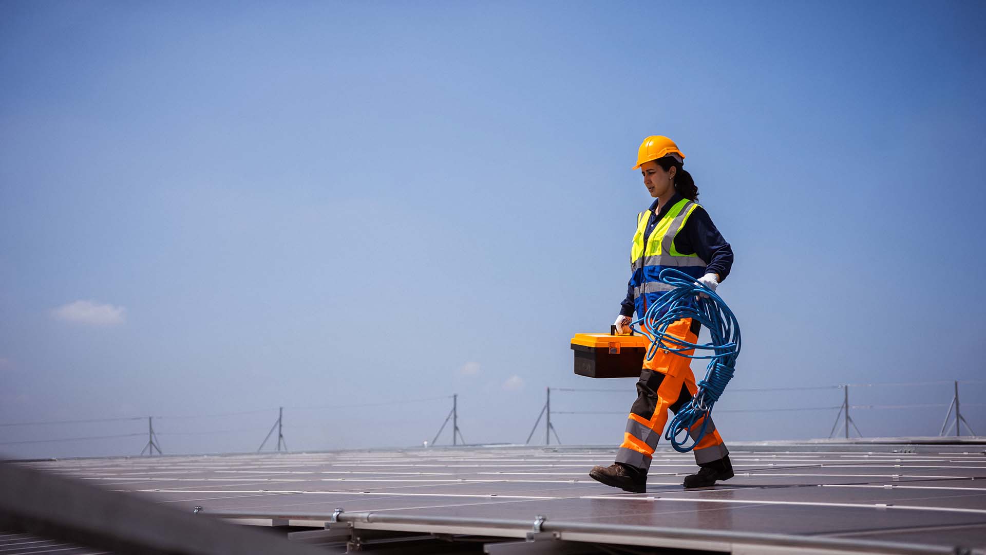 Woman wearing safety equipment and carrying maintenance equipment walks across solar panels. 