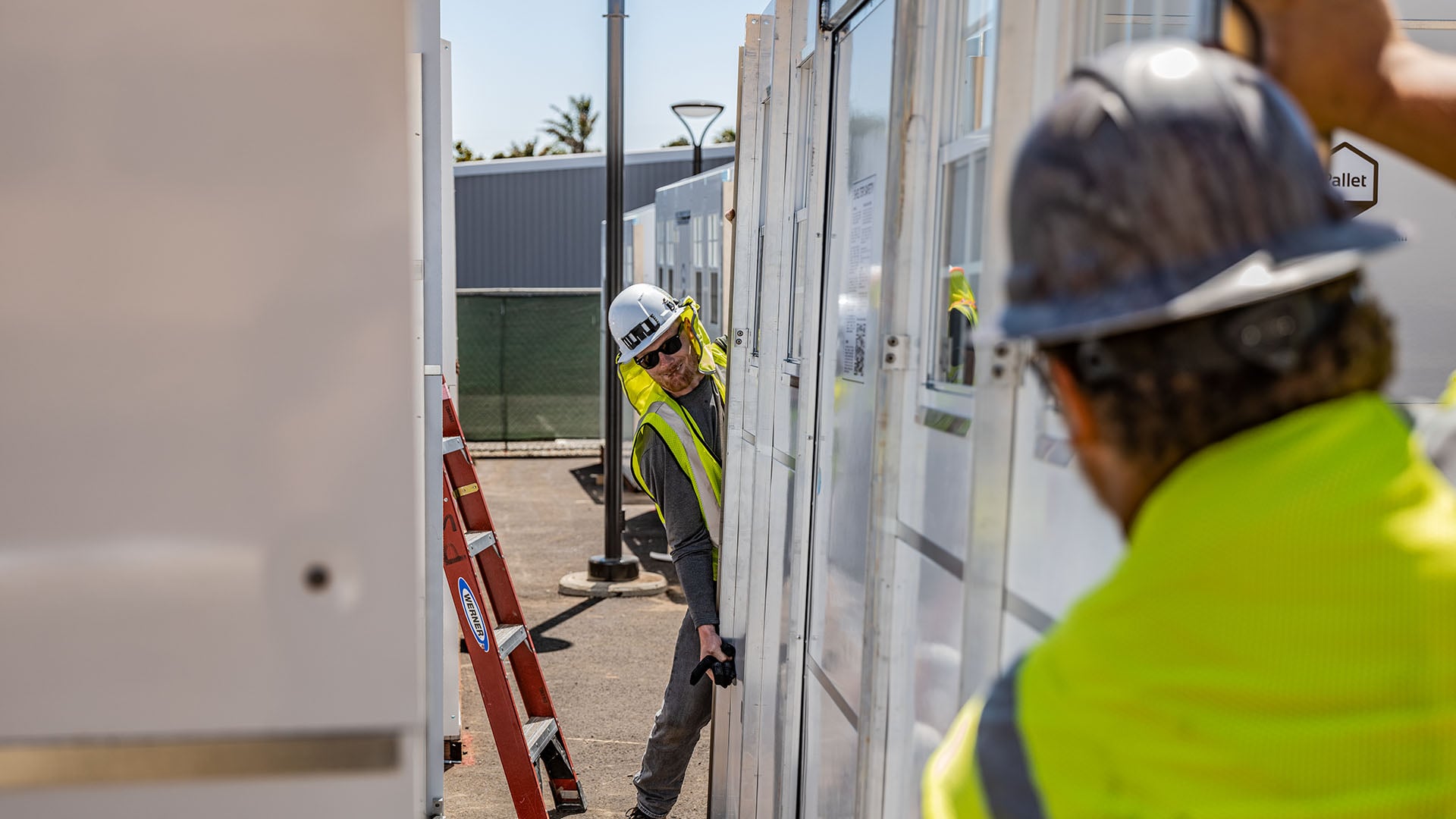Two men wearing high vis vests and hard hats work together to lift shed. 