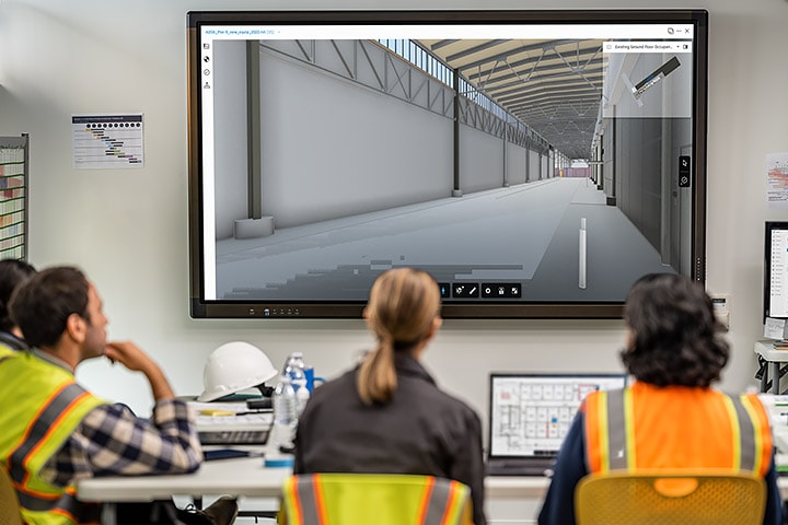 Five people in construction-industry garb seated at a table and viewing plans on a large digital wall screen 
