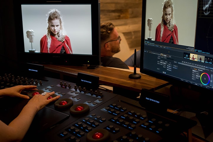 A visual artist seated at a console making digital color edits to an image of a woman in a red cape 