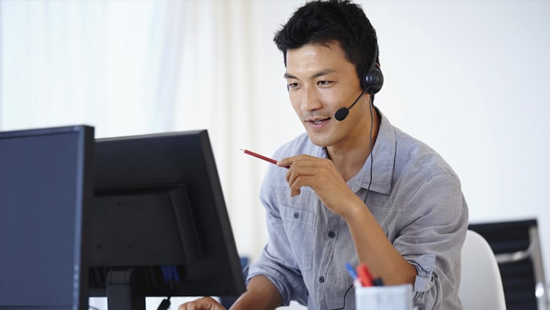Een man in een kantoor kijkt naar zijn desktopscherm terwijl hij praat door een hoofdtelefoon.  