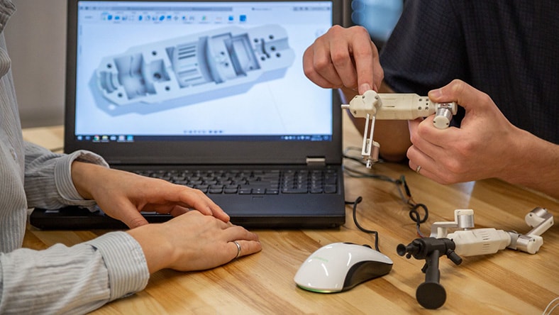 Two people assembling a 3D-printed mechanical component while referencing a CAD design displayed on a laptop screen.