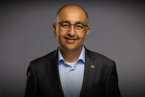 Smiling middle-aged man wearing glasses, a dark suit jacket, and a light blue shirt against a plain gray background