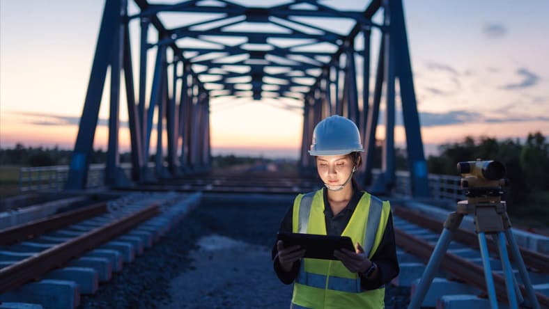 Ramboll engineer in a hard hat and hi-vis vest using a tablet on railway tracks under a steel bridge, showing better project delivery